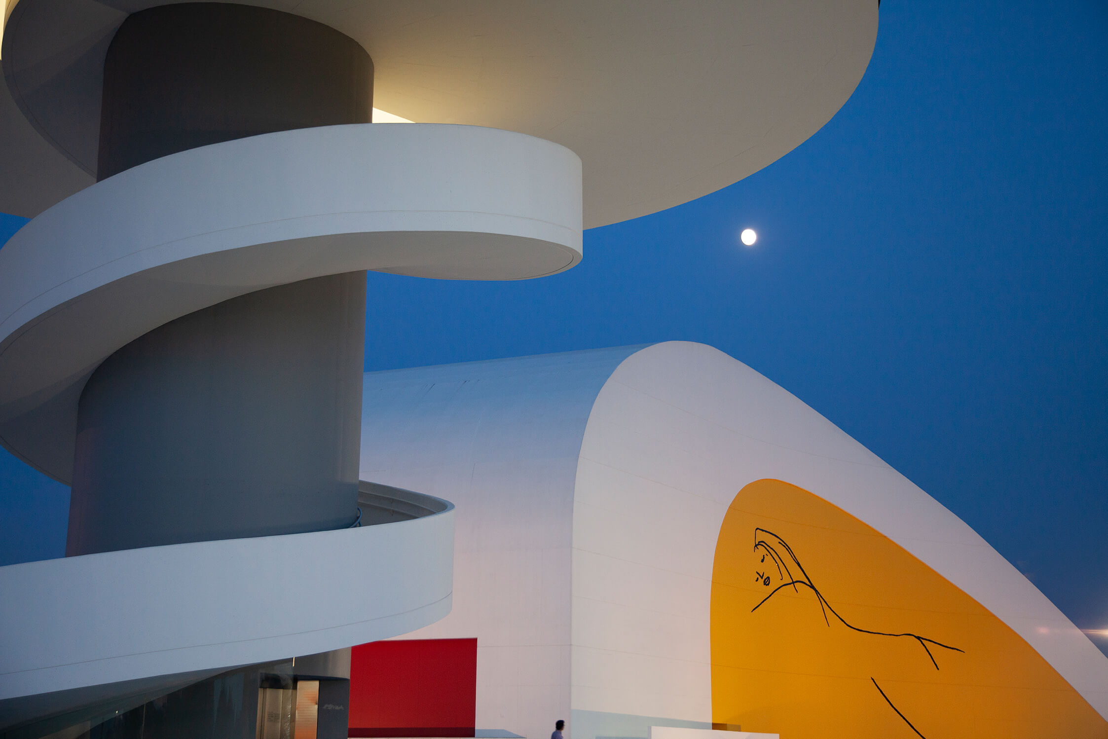 Un primer plano de una gran escalera de caracol ocupa la mitad izquierda de la foto, a su lado un edificio de formas curvas y arriba aparece la luna casi llena.